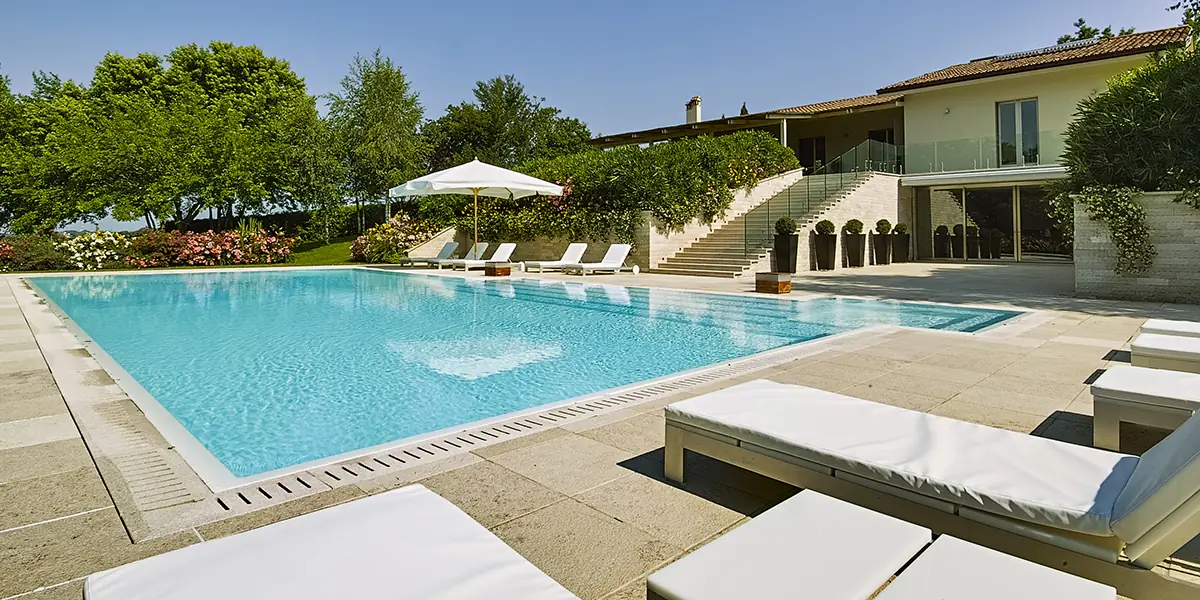 Luxurious swimming pool surrounded by sun loungers, lush greenery, and a modern home, showcasing ideal conditions for pool resurfacing services by Classic Marcite in Jacksonville Beach.