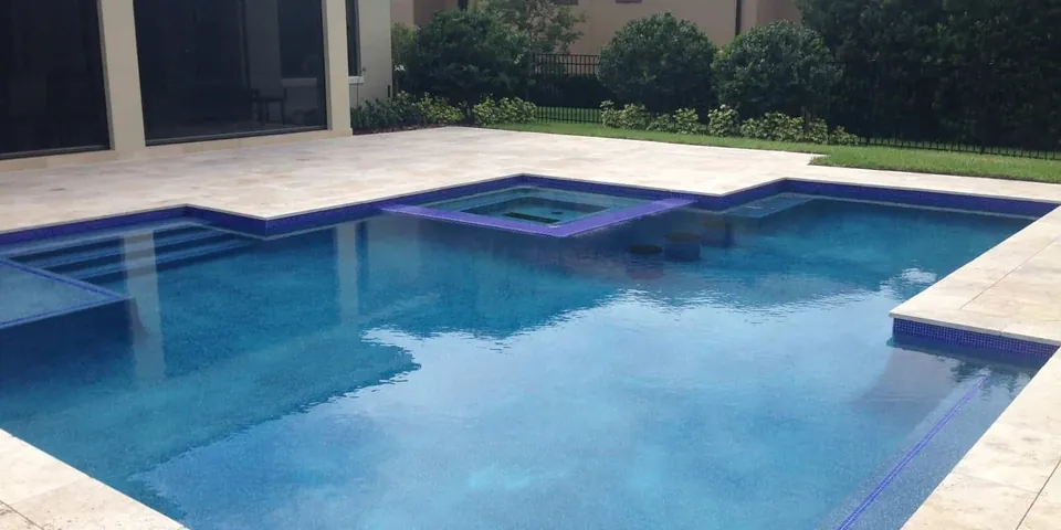 Pool and spa with travertine deck and purple tile beside a home