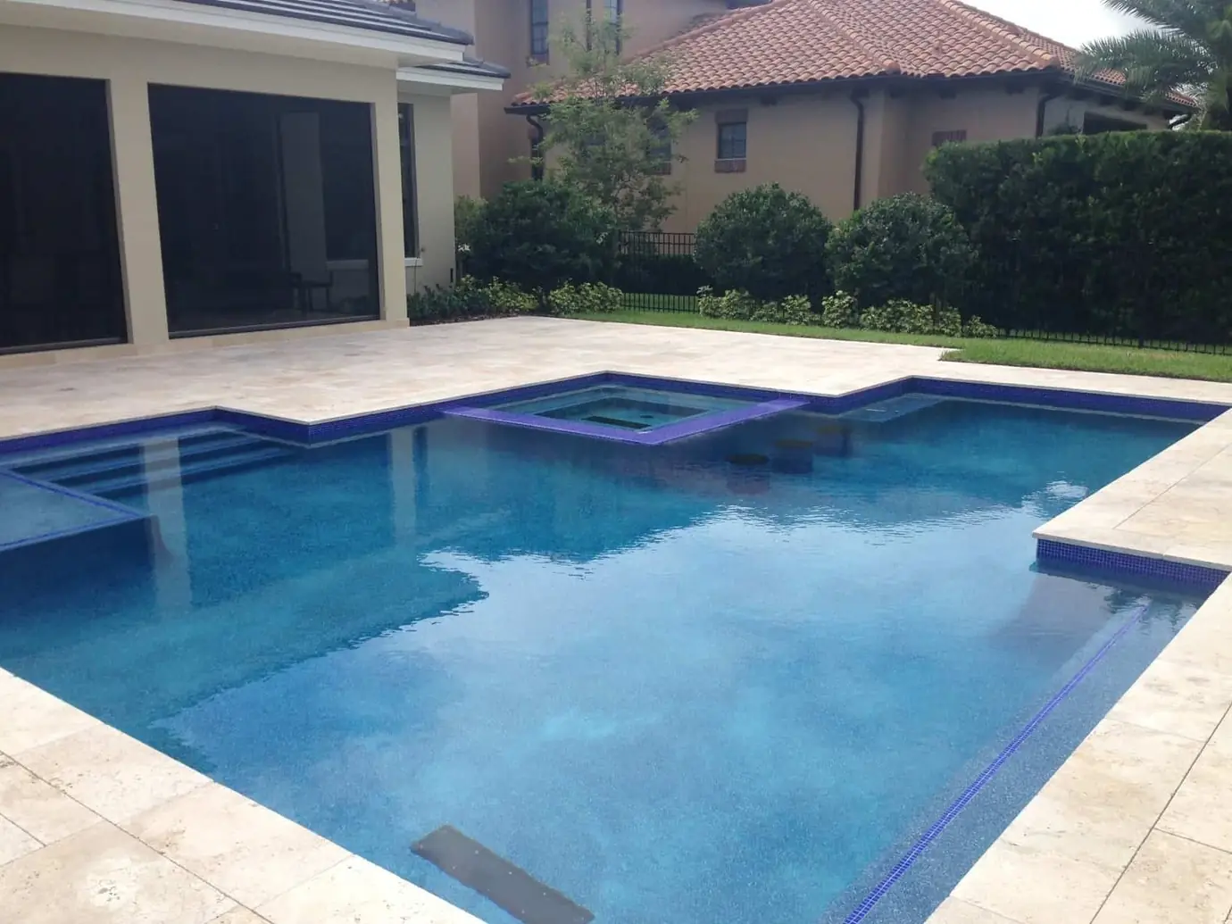 Pool and spa with travertine deck and purple tile beside a home